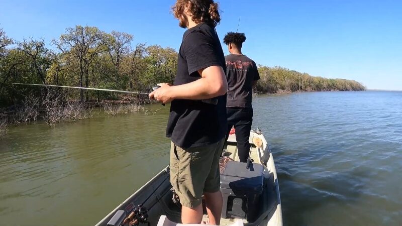 The Image Shows Two Individuals Standing on A Small Boat, Fishing in The Calm Waters of Lake Arlington Near a Shoreline Lined with Trees, Enjoying a Peaceful Day of Boating and Fishing Under Clear Blue Skies