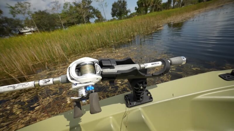 A Close-Up View of A Fishing Rod and Reel Mounted on A Kayak, Positioned Near Reeds on The Edge of A Serene Lake