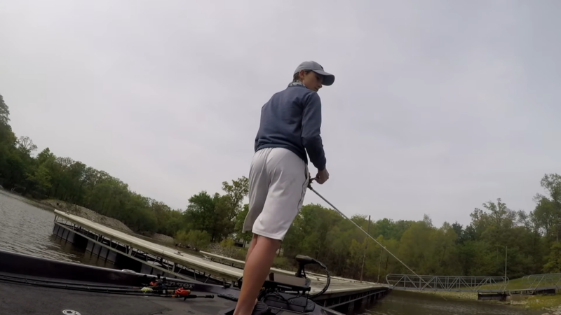 The Image Shows a Person Fishing While Standing on A Boat Near a Dock at Carlyle Lake, with Trees and A Cloudy Sky in The Background