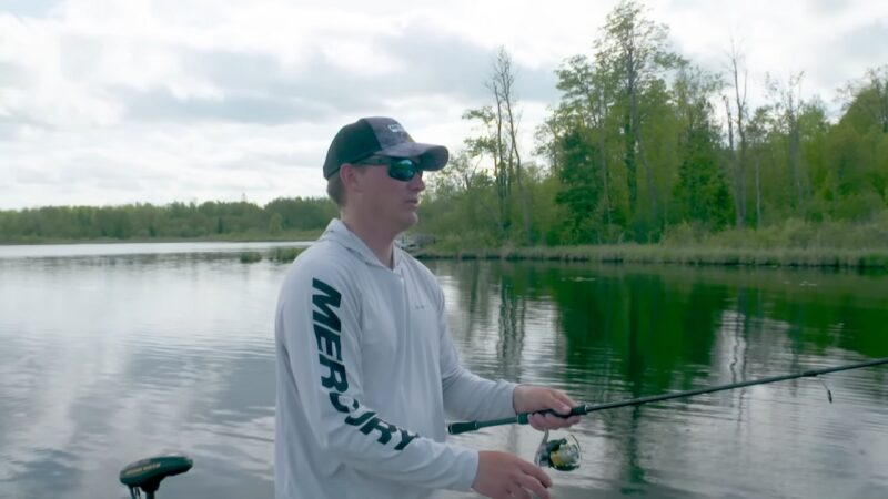 A Man in A Mercury-Branded Shirt Fishes from A Boat, Focusing Intently on His Line in A Calm, Tree-Lined Lake