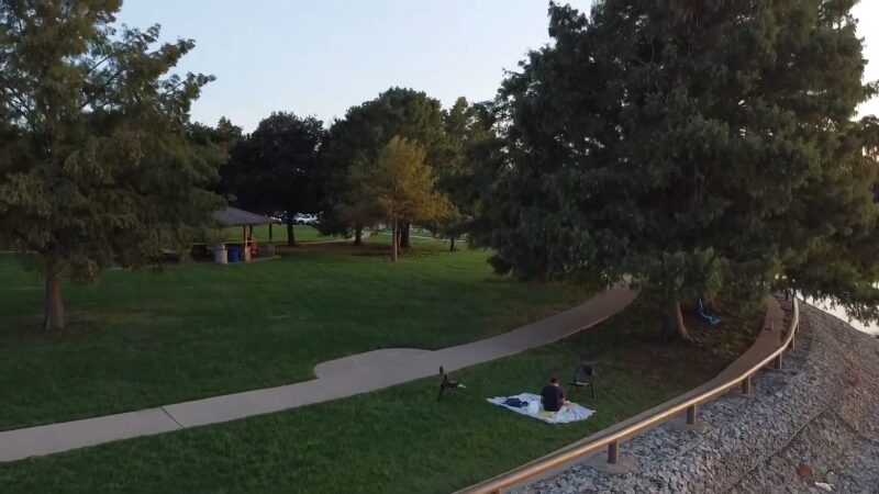 The Image Shows a Peaceful Picnic Area at Lake Arlington, Featuring a Person Sitting on A Blanket by A Tree-Lined Path with A Nearby Pavilion, Surrounded by Green Grass and Trees Near the Water's Edge