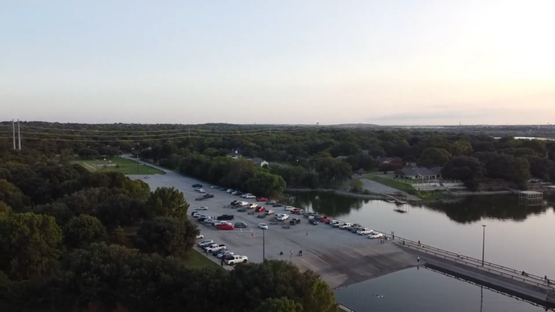 The Image Shows a Parking Lot Near a Boat Ramp at Lake Arlington, Surrounded by Trees and Calm Water, with A Few People and Cars, Indicating a Well-Organized Recreational Area During the Early Evening