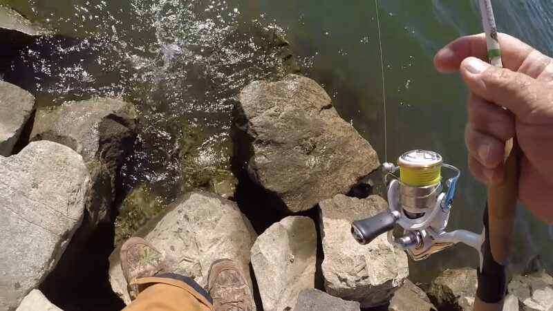 A Fisherman Holding a Fishing Rod with A Spinning Reel, Standing on Rocks by A River