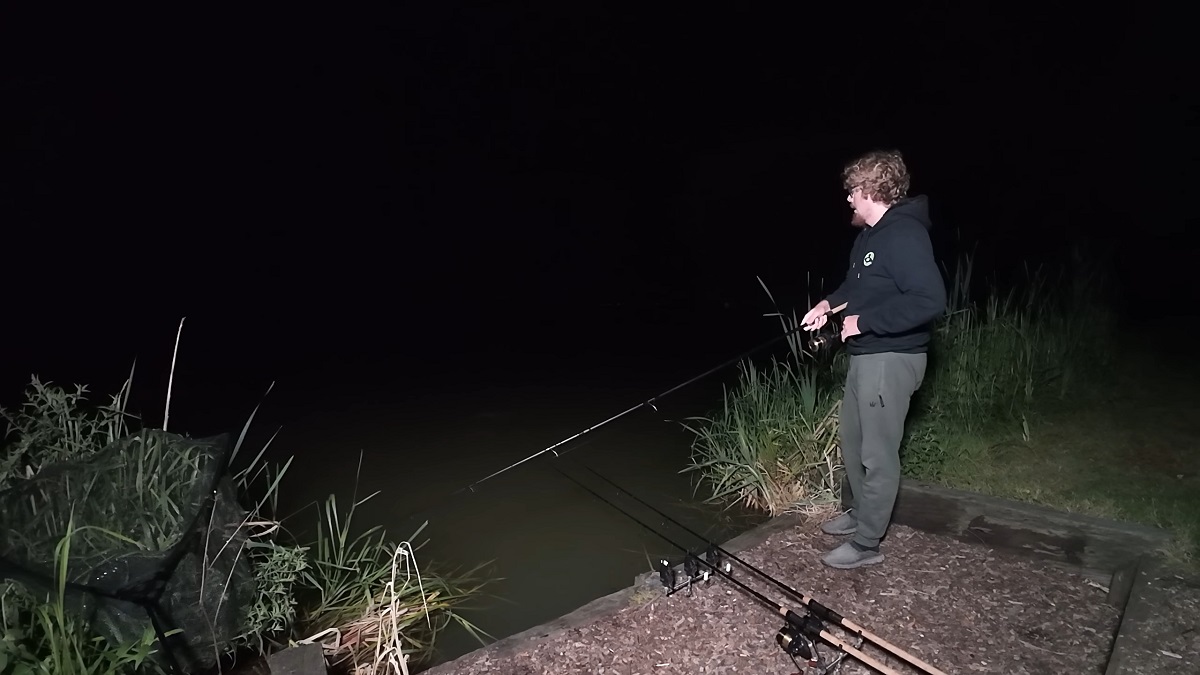 A man standing by the water at night, holding a fishing rod while focusing on his line